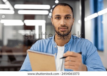 Close-up portrait of a young hispanic man sitting in a modern office, holding a pen and notebook and looking confidently at the camera.