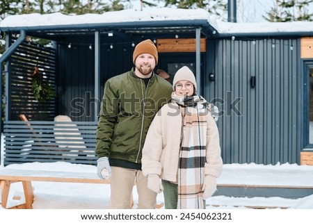 Medium long portrait of happy Caucasian man and his daughter in casual warm clothes standing outdoors against their cottage on winter day, copy space