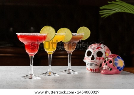 An array of margarita cocktails in different flavors, garnished with lime, beside vibrant sugar skulls, symbolizing the Mexican Day of the Dead