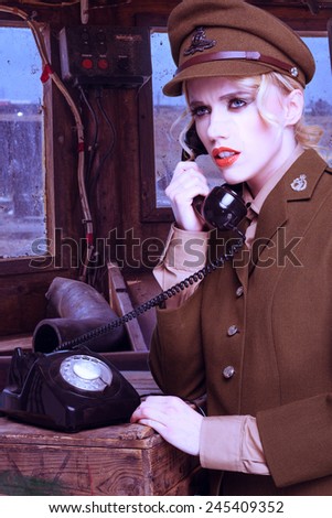 Pretty blond woman in a brown army uniform standing talking on a retro dial-up telephone on a wooden crate in a barracks