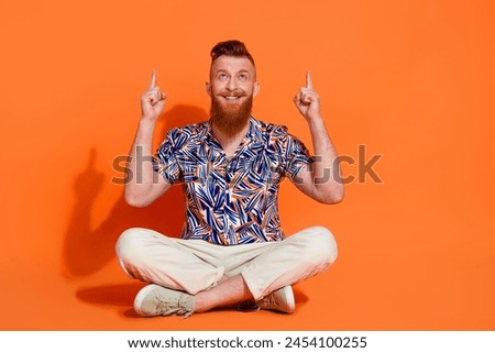 Full size photo of good mood man wear stylish shirt sit on floor look directing at logo empty space isolated on orange color background