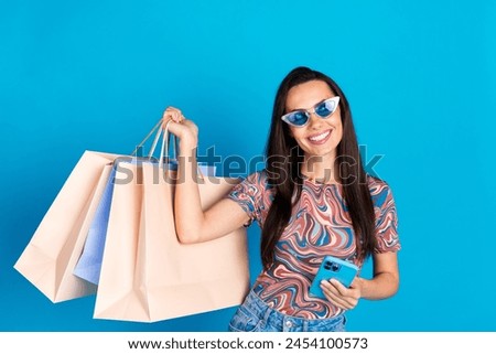 Photo of pretty young girl sunglass hold device shopper look empty space wear trendy colorful clothes isolated on blue color background