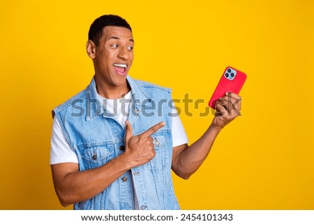 Portrait of multiethnic multinational man dressed denim vest over t-shirt directing look at smartphone isolated on yellow color background Royalty-Free Stock Photo #2454101343