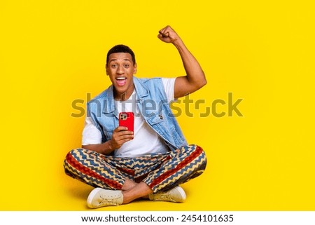 Full size photo of overjoyed man dressed denim vest sit on floor holding smartphone win lottery isolated on yellow color background