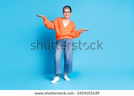 Full body photo of lovely senior lady compare palm hold empty space dressed stylish orange garment isolated on blue color background