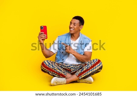 Full size photo of clever good mood man dressed denim vest sit on floor look directing at smartphone isolated on yellow color background