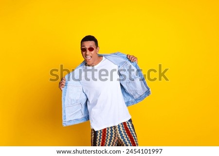 Full size photo of cool eccentric man dressed denim vest over t-shirt in sunglass holding jacket isolated on yellow color background