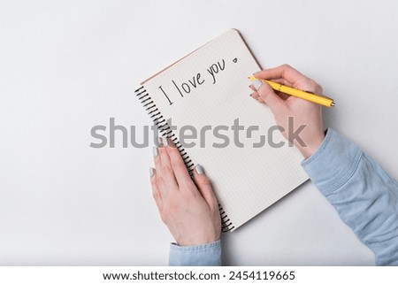 Female hand writing in notebook the words I LOVE YOU. Top view of notebook and hands. White background.
