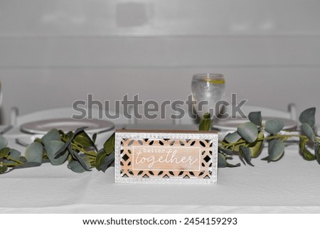 Table setting with a decorative 'better together' sign, eucalyptus garland, and elegant dinnerware. Wedding reception and romantic event decor concept.