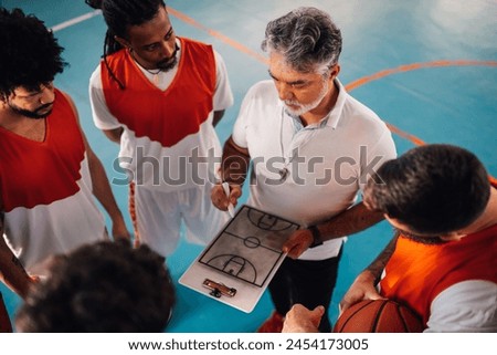 High angle view of a mature basketball coach and his diverse team having a team talk while gathered together at the court and cheering each other. Elderly coach drawing defense on a clipboard.