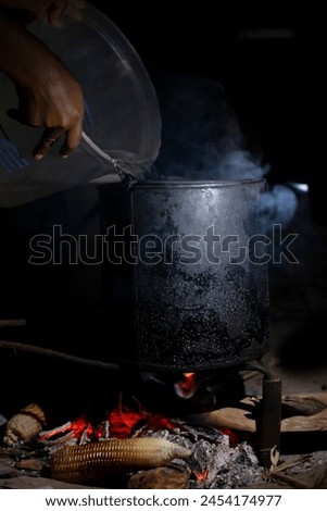 A picture of water being pored in a water boiling can and corn being grilled in a fire place.