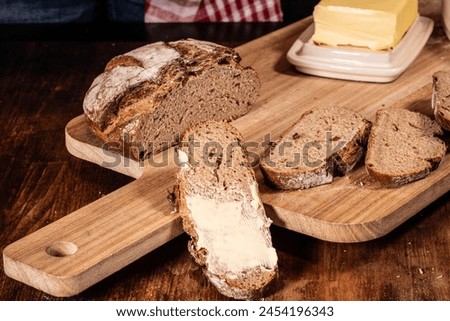 
Sliced artisanal bread on a wooden chopping board with a pat of butter, captured in the midst of spreading, embodies homemade comfort and simple culinary delights. Royalty-Free Stock Photo #2454196343