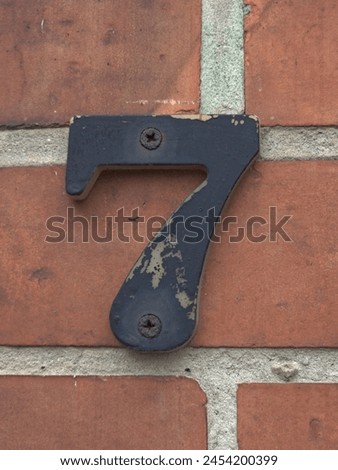 A sign with the number seven (7) on the house, fastened with rusty screws. Against the background of a brick wall. Black peeling paint