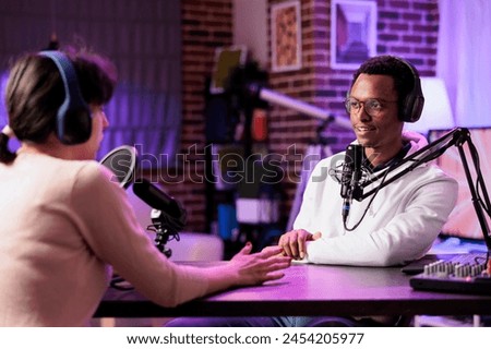 Man and woman broadcasting discussion on online podcast episode using professional equipment . Vlogger show host and guest talking in neon lights ornate living room personal studio broadcast Royalty-Free Stock Photo #2454205977