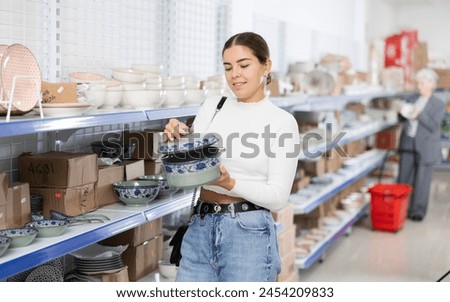Smiling interested young girl making purchases in Asian store, choosing traditional ceramic soup tureen.. Royalty-Free Stock Photo #2454209833