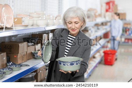 Positive senior female shopper choosing with interest oriental style porcelain soup tureen among variety of dishware arranged on shelves at Asian store Royalty-Free Stock Photo #2454210119