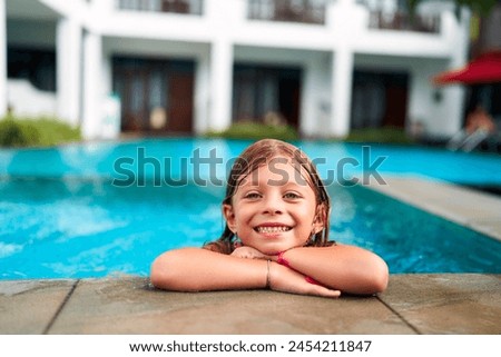 Smiling girl enjoys summer fun in resort pool. Child with wet hair leans on edge, happy in water. Bright day for kids outdoor swimming activity. Playful moment, family vacation joy at tropical hotel.