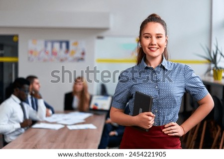 Confident asian businesswoman holds tablet in modern office, diverse team working in background. Pro in smart casual attire, ready for corporate strategy session. Workplace equality focus.