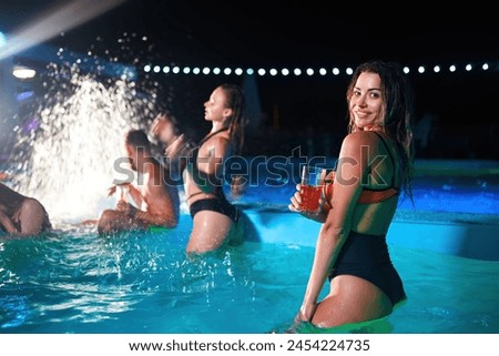 Pretty woman in bikini drinking cocktail and hanging out with friends at night pool party. People having fun dancing with drinks and splashing water in luxury private villa swimming pool.