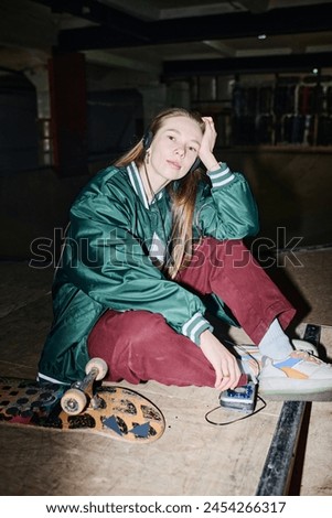 Vertical flash shot of young Caucasian woman in streetwear sitting on floor in skatepark listening to music on retro portable player