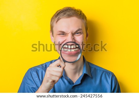Young man close-up in a blue shirt on a yellow background holding a magnifying glass, looking into it. Funny/ Mimicry. Gesture. photo Shoot