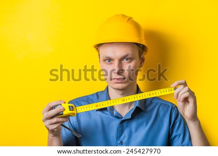 Young man close-up in a blue shirt and construction helmet on a yellow background holding a tape measure, measure something. Mimicry. Gesture. photo Shoot