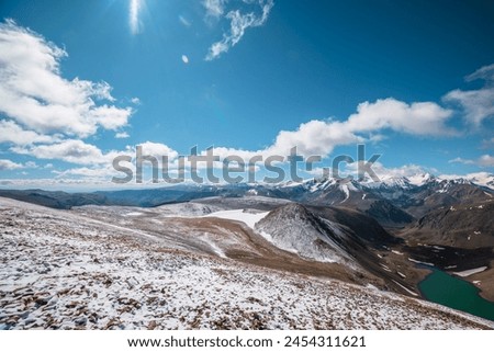 High snowy stony pass with view to most beautiful rocky hill in freshly fallen snow, alpine lake and large colorful mountain range with snow-white peaks in far away in low clouds in sunny cloudy day. Royalty-Free Stock Photo #2454311621