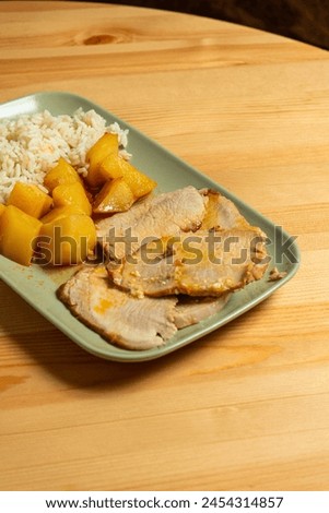 A succulent roasted pork loin sits on a plate alongside golden-brown roasted potatoes and a mound of fluffy white rice. The dish is presented in a cozy restaurant setting, inviting and warm