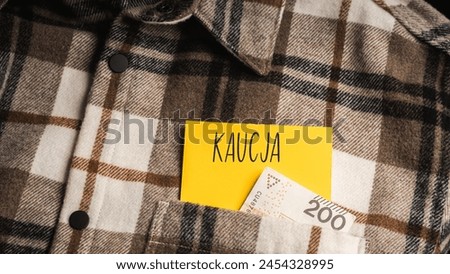 Yellow card with a handwritten inscription "Kaucja", protruding from a brown plaid shirt, next to Polish banknotes PLN (selective focus), translation: deposit
