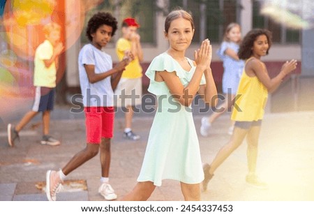 Happy cute girl dancing modern choreography with group of tweenagers on city street on sunny summer day, toned image..