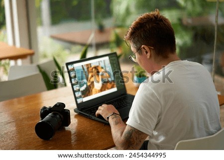 Focused photo editor with tattoos retouches images on laptop at coworking space. Professional with camera reviews creative visual content. Freelancer works in modern office, digital workflow.