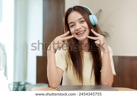 Asian woman with headphones and braces makes peace sign with fingers, at home in front of a laptop, emanating cheerfulness. Energetic individual signals V-sign, orthodontic braces on show