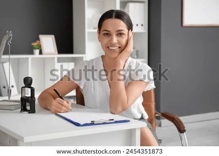 Pretty businesswoman sitting at table and writing on clipboard in light office
