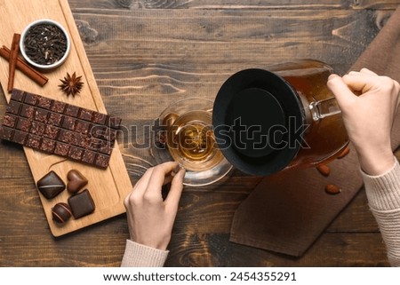 Woman pouring tasty hot tea from teapot into cup and tray with chocolate on wooden background
