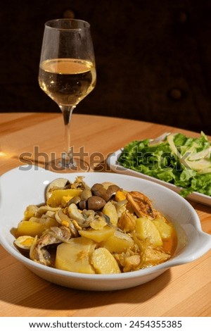 A dish of Bacalhau à Gomes de Sá with baked cod, potatoes, and onions, topped with black olives and hard-boiled eggs. Paired with a glass of white Alentejo wine and a lettuce salad in a restaurant.