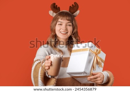 Pretty young woman holding cup of hot chocolate with marshmallows and Christmas gift box on orange background