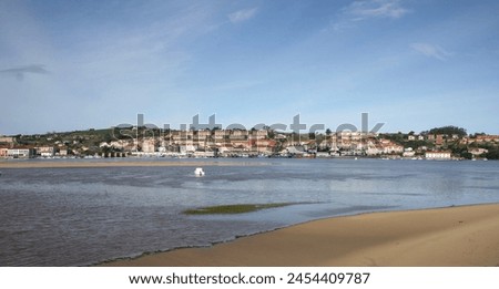 Picture of San Vicente de la Barquera on a clear and calm day where you can see the boats moored in the bay at low tide and in the background the mountains.
