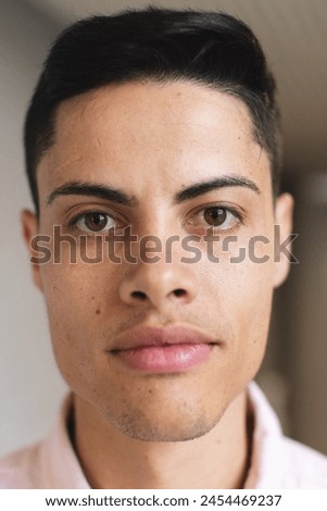 A biracial young man with short dark hair is looking directly at the camera in a modern business office. He has light brown skin, brown eyes, and is wearing a pink shirt, unaltered.