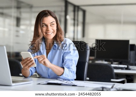 Busy happy middle aged business woman holding cell mobile phone using cellphone looking away in office. Smiling mature businesswoman executive professional leader working on smartphone. Copy space.