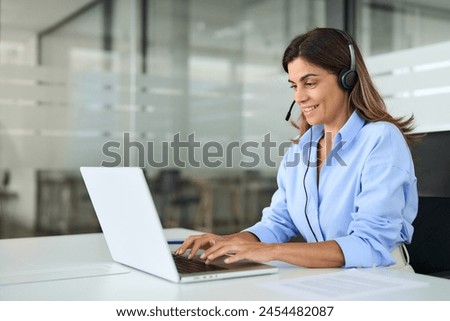 Busy mature business woman call center representative customer support agent helping client, smiling middle aged senior female operator wearing headset working using laptop computer in office. Royalty-Free Stock Photo #2454482087