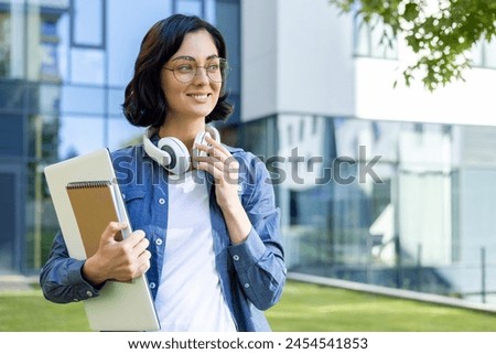 A young smiling female student stands on the street in headphones, holds a laptop and notebooks in her hand, looks to the side.