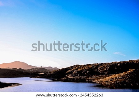 A beautiful mountain range with a calm river running through it. The sky is clear and blue, and the sun is setting, creating a warm and peaceful atmosphere