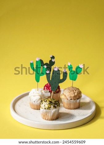 This is an image of a playful and creative dessert display. There are three cupcakes arranged on a white plate, each with a different topping, and they are decorated to resemble cacti. The cacti are m