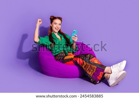 Full size photo of overjoyed woman dressed green shirt sit on bean bag hold smartphone win lottery isolated on purple color background