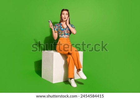 Full size photo of pretty young woman sit cube hold phone empty space wear top isolated on bright green color background