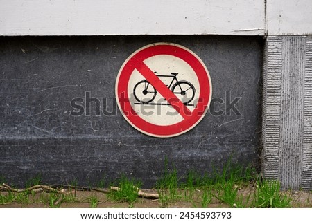 Bike bicycle symbol icon forbidding parking storing leaving on curb sidewalk pavement sign red white and black warning on building cement wood grass growing out of concrete cracks city street road