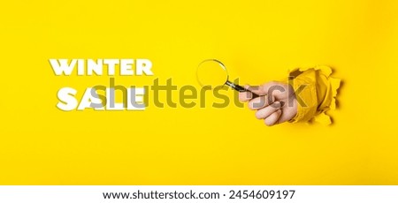 A hand holding a magnifying glass with the words Winter Sale written below it. The image has a playful and lighthearted mood