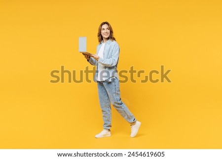Full body smiling young IT woman she wear blue shirt white t-shirt casual clothes hold use work on laptop pc computer look aside isolated on plain yellow background studio portrait. Lifestyle concept