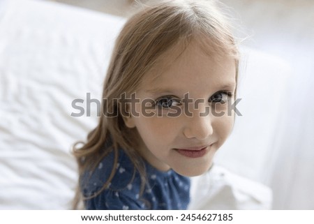 Pretty little awakened girl looking at camera seated on white sheets on bed, having attractive appearance, happy mood and curious character explore, watching at you, close up. Eye sight, vision care