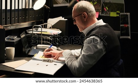 Law agent takes notes on files, writing clues from classified records to connect the dots and solve criminal case. Investigator working in incident room, reading confidential papers. Handheld shot. Royalty-Free Stock Photo #2454638365